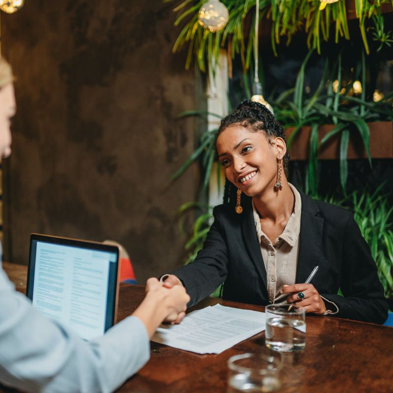 Two people shaking hands at a table with a laptop and documents in an indoor setting.